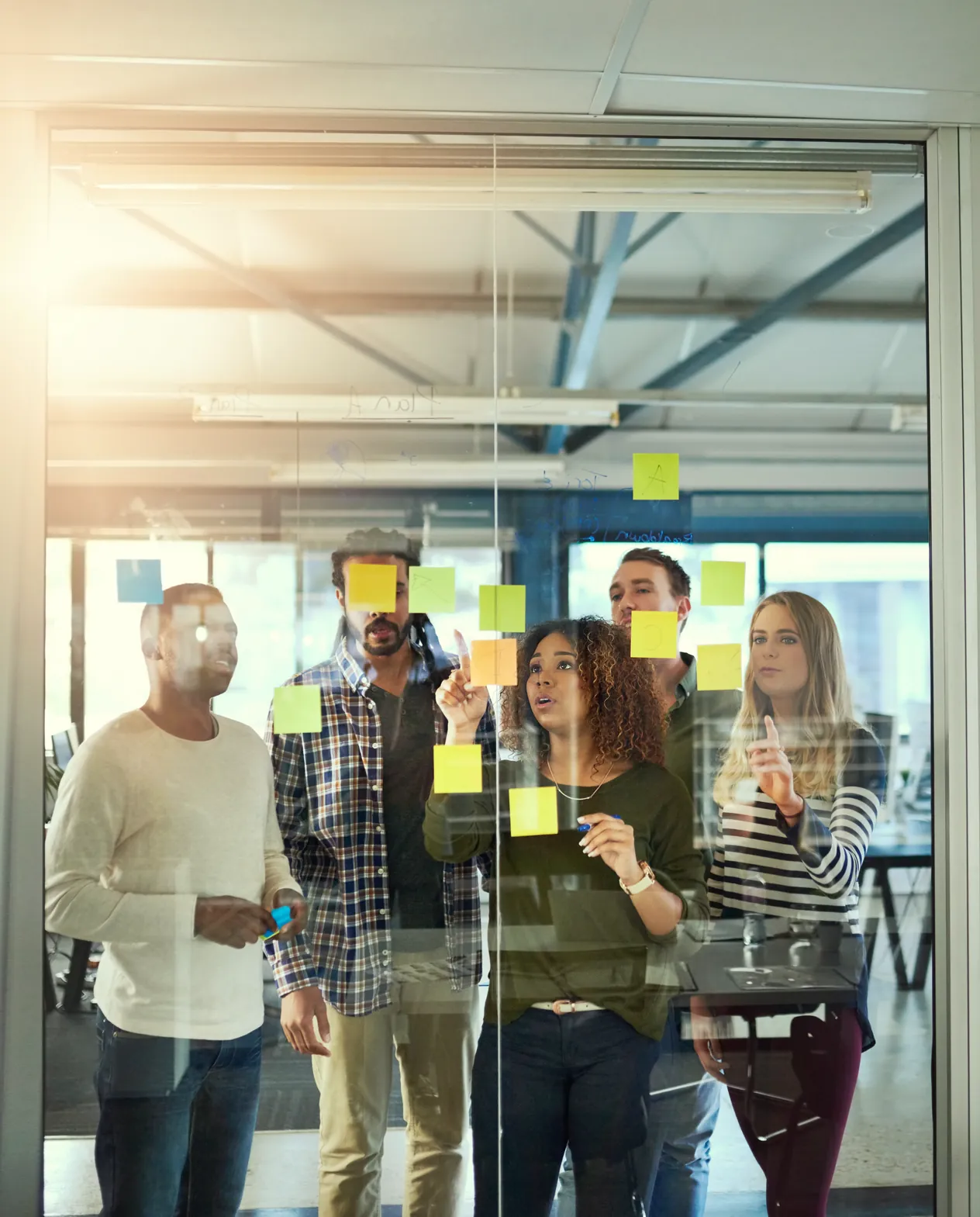 Five colleagues seen through a glass wall covered in sticky notes, adding another to it from the far side