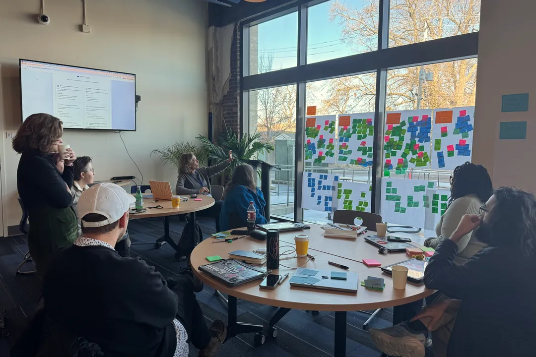 Teachers around a table watching a screen, sticky-note boards filling the windows behind them