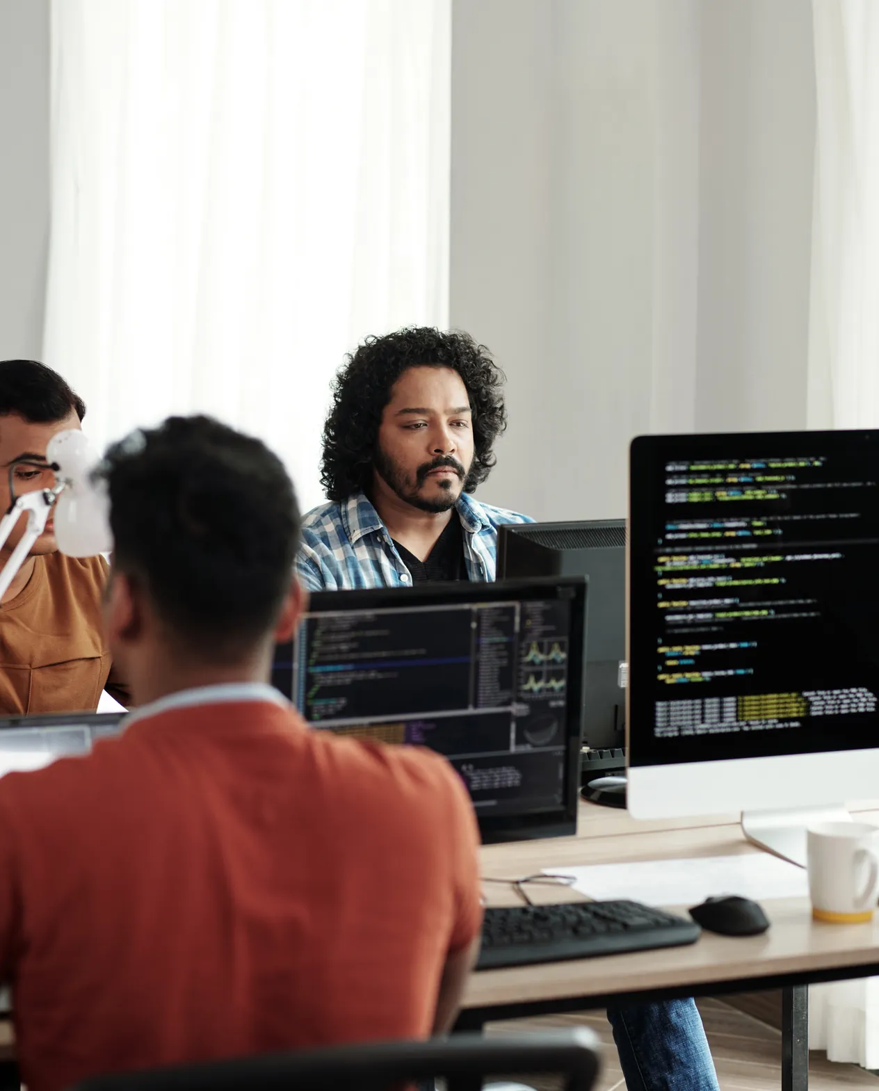 Two engineers at adjacent desks, one of them reading lines of code on the monitor in front of him