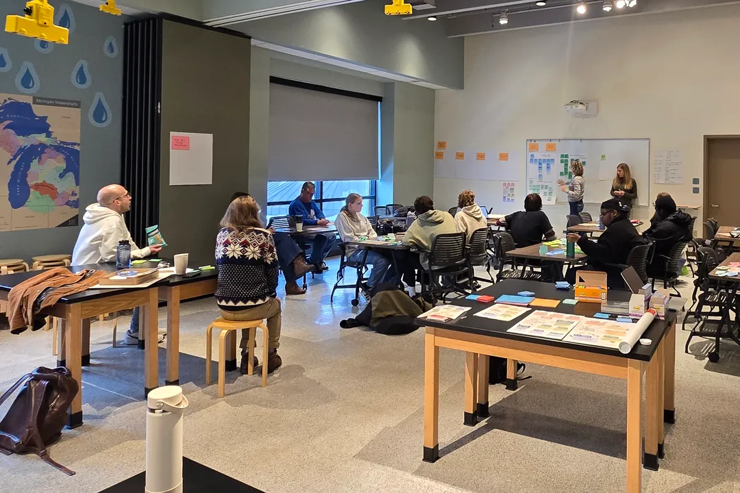 Teachers at worktables in a high-ceilinged classroom, project cards and sticky notes spread in front of them
