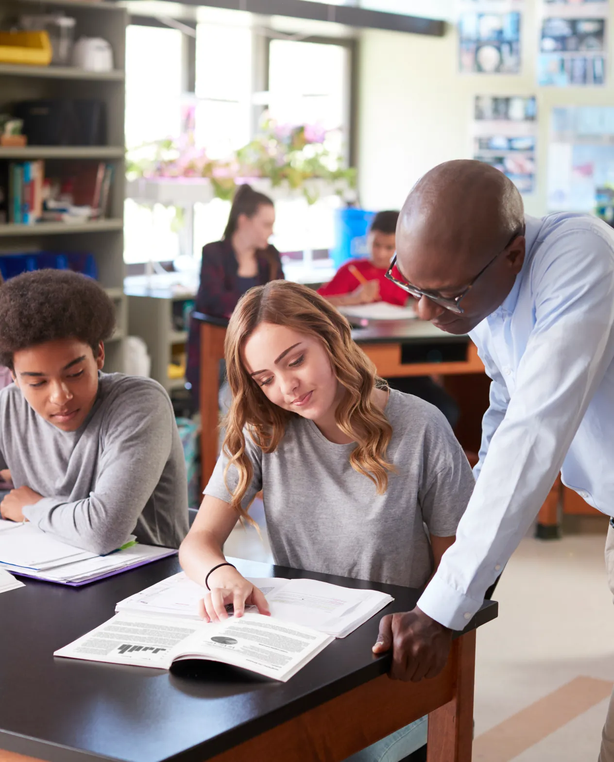 A teacher leaning over a student’s desk to read an open textbook with her, another student working alongside