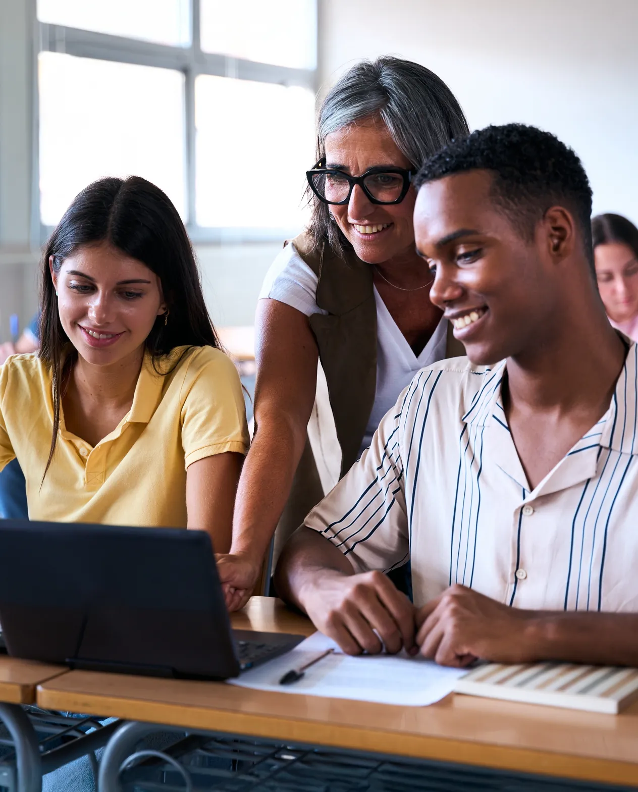 A teacher leaning between two students at a desk, all three looking at the laptop in front of them